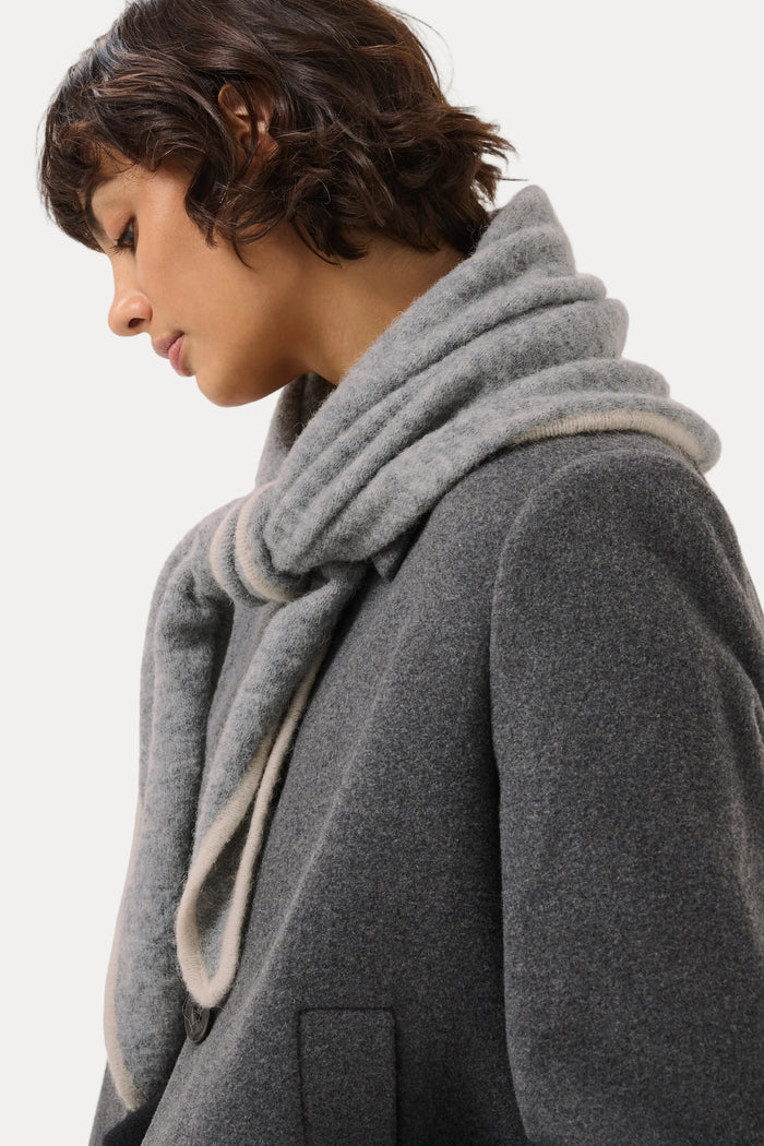 A close-up side profile of a person wearing a thick, double-layered gray knit scarf and a dark gray wool coat against a plain light gray background.