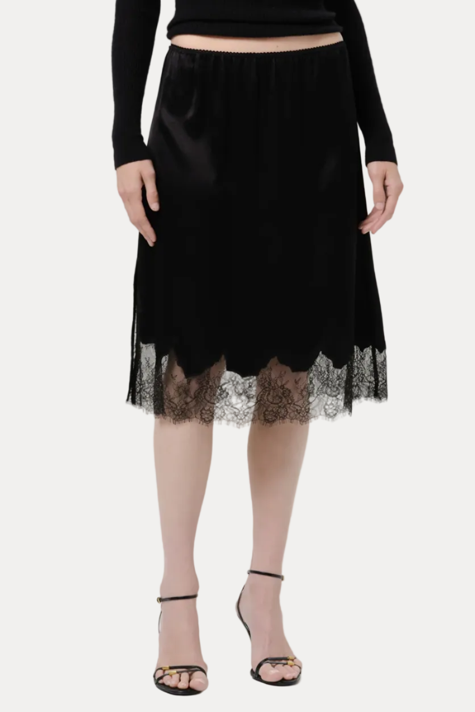 A model wearing a black velvet midi skirt with a scalloped black lace hem, paired with a black long-sleeve top and black strappy heels, against a light gray studio background.