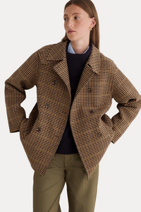 A woman wearing a brown and beige houndstooth double-breasted blazer over a dark navy sweater and olive green trousers, posing against a light gray wall.