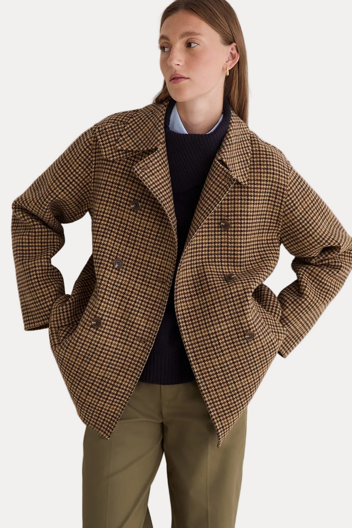 A woman wearing a brown and beige houndstooth double-breasted blazer over a dark navy sweater and olive green trousers, posing against a light gray wall.