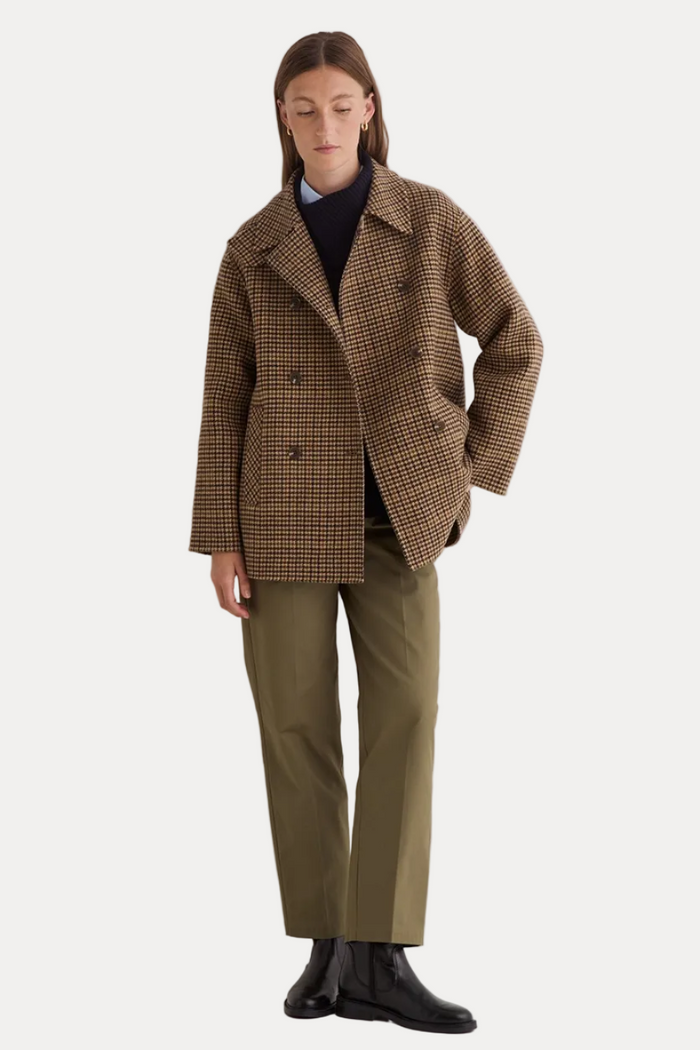 A model stands against a plain white studio wall wearing a brown and beige houndstooth double-breasted blazer, olive green trousers, a black turtleneck, and black leather boots.