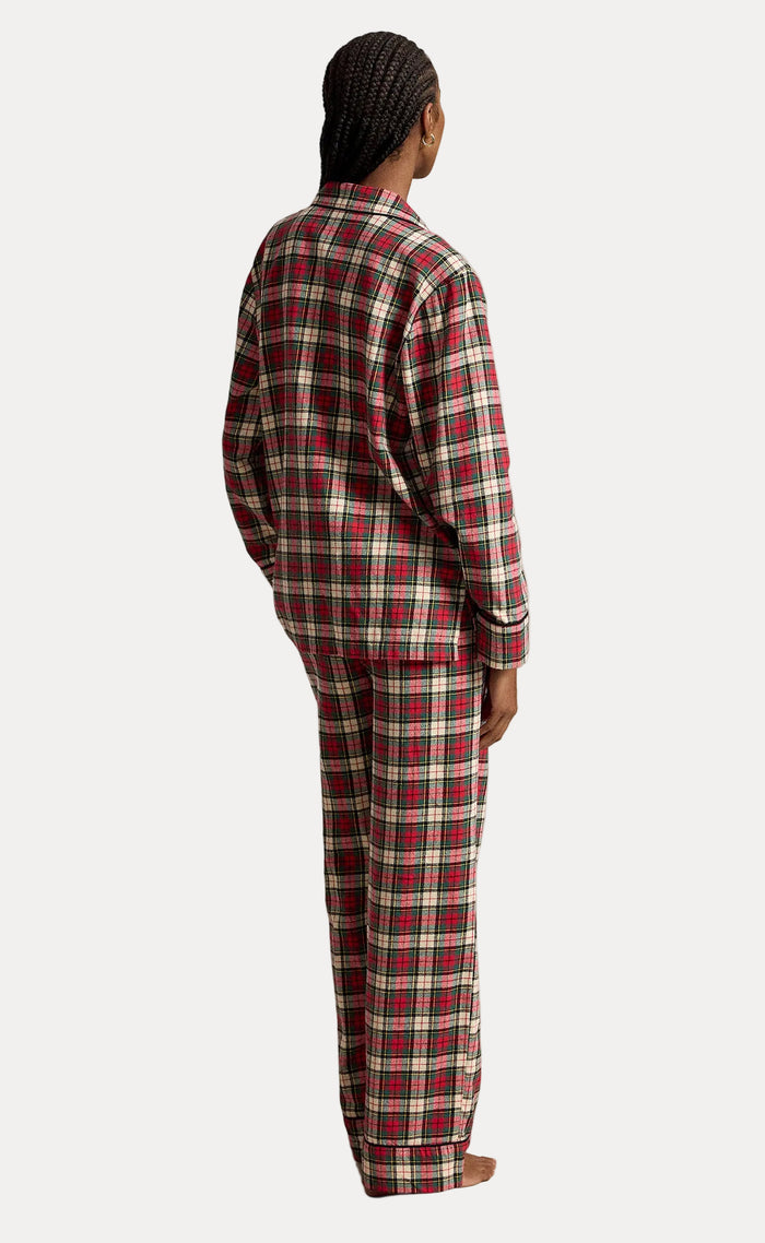 A person shown from the back wearing a matching long-sleeve plaid pajama set in red, beige, and green, standing against a neutral textured studio backdrop.