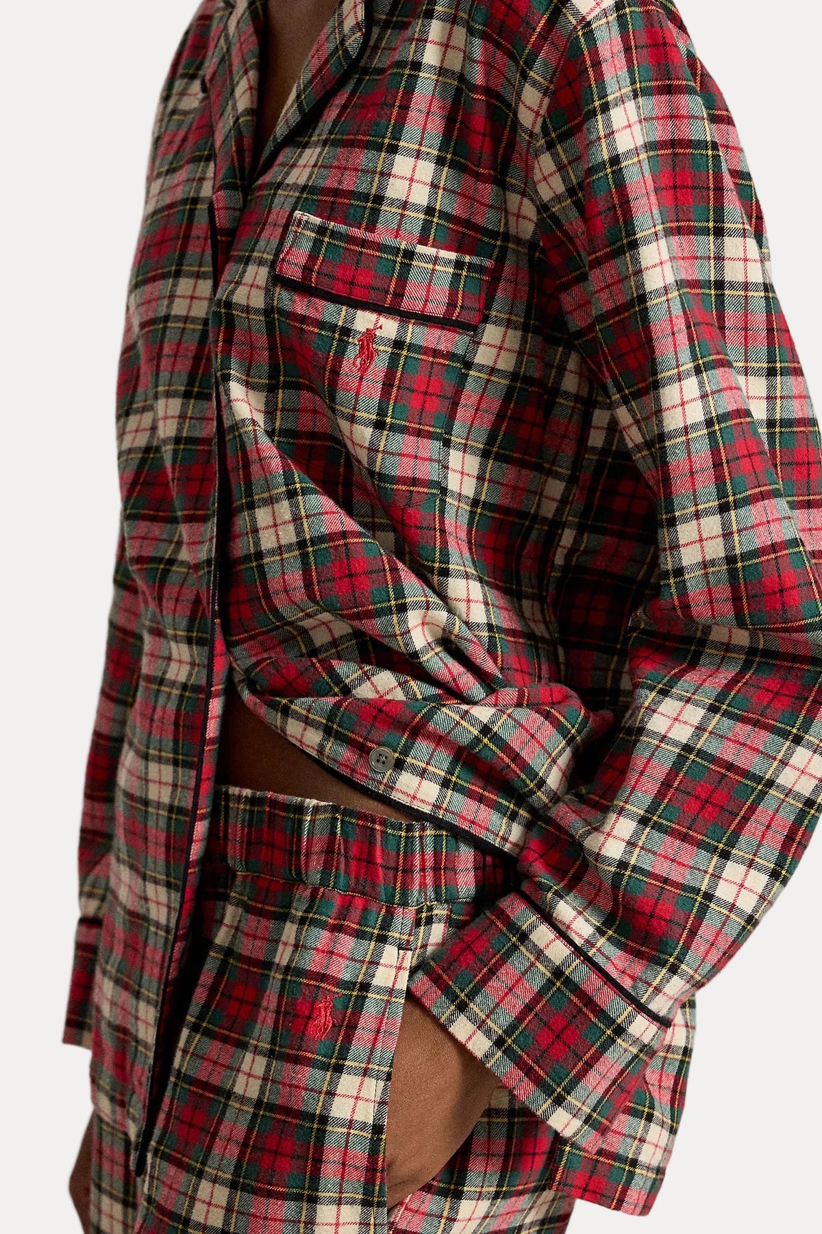 A close-up of a person wearing a red, green, and cream plaid button-up shirt and matching plaid shorts, with one sleeve rolled up and a hand in the pocket.