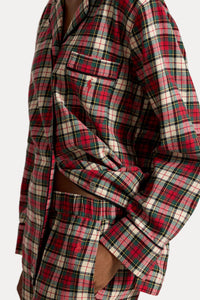 A close-up of a person wearing a red, green, and cream plaid button-up shirt and matching plaid shorts, with one sleeve rolled up and a hand in the pocket.