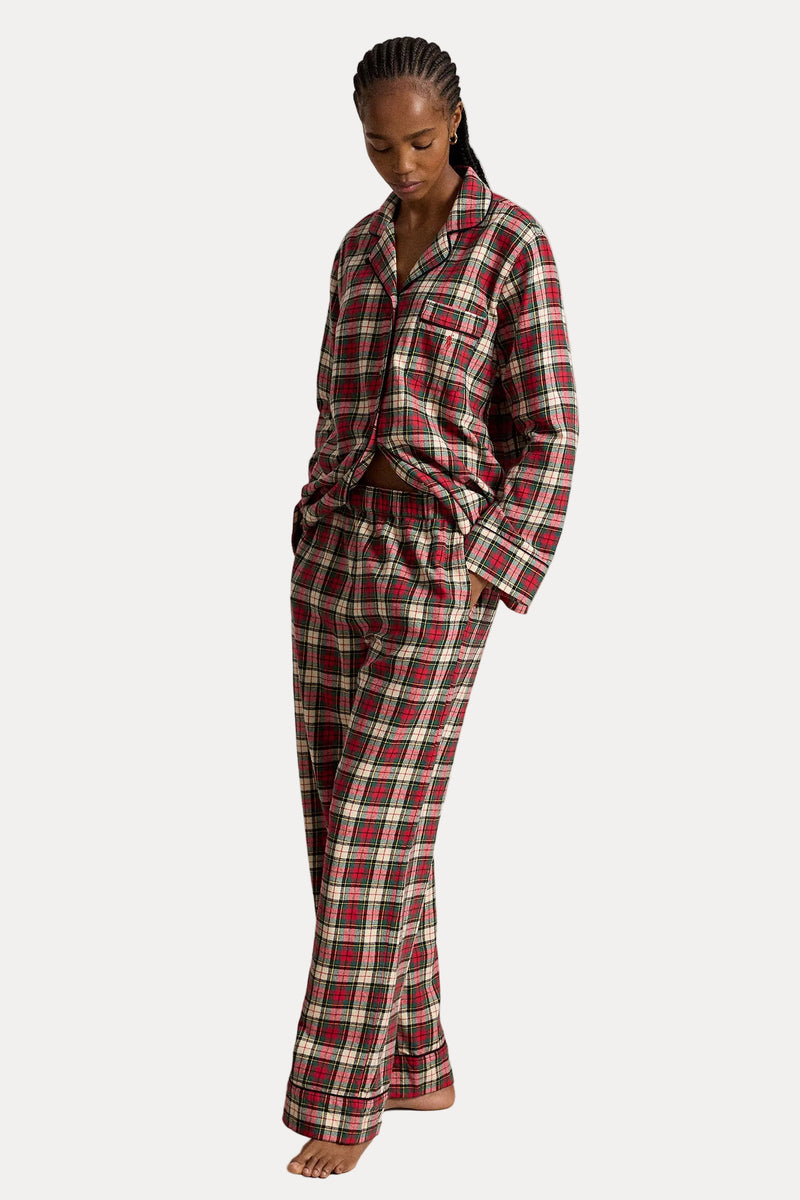 A woman modeling a red, green, and cream plaid pajama set consisting of a long-sleeve button-up shirt and matching pants, standing against a neutral beige studio backdrop.