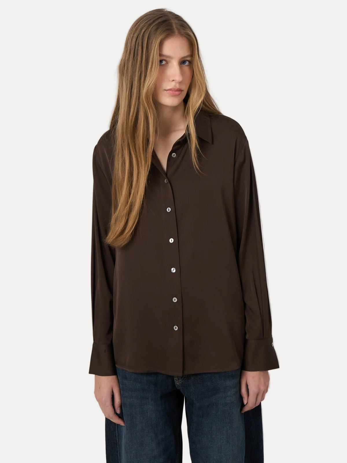 A woman modeling a dark brown button-up shirt with a pointed collar and long sleeves, paired with dark blue jeans, against a plain light gray background.