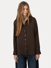 A woman modeling a dark brown button-up shirt with a pointed collar and long sleeves, paired with dark blue jeans, against a plain light gray background.