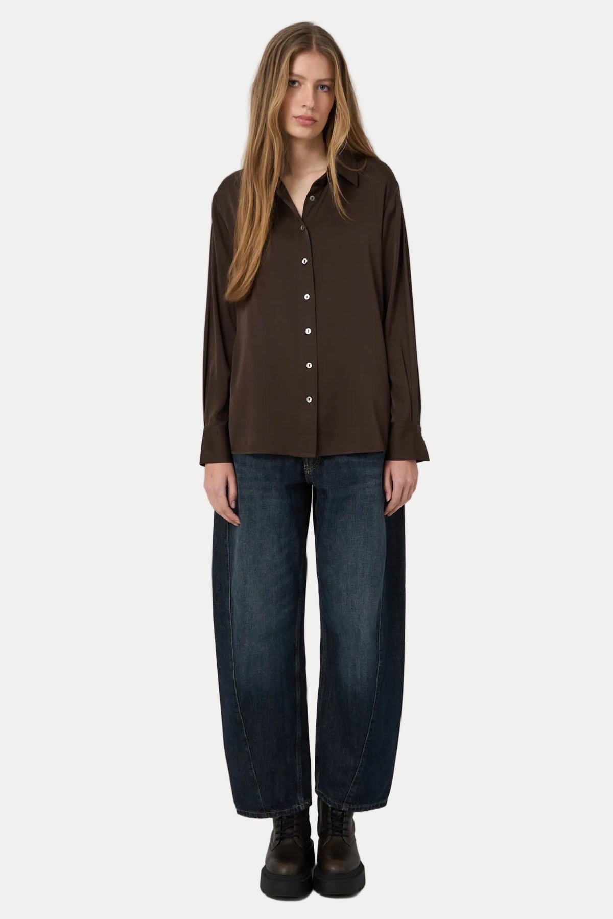 A woman stands against a plain light gray studio background wearing a dark brown long-sleeve button-front shirt, loose dark blue jeans, and dark brown boots.
