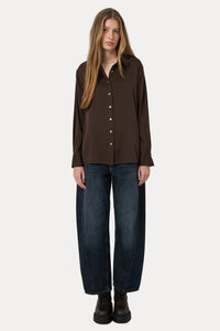 A woman stands against a plain light gray studio background wearing a dark brown long-sleeve button-front shirt, loose dark blue jeans, and dark brown boots.