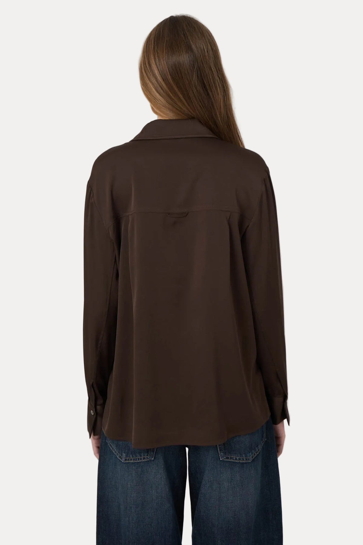 A woman viewed from behind wearing a dark brown long-sleeve button-up shirt and blue jeans against a plain light gray background.