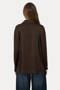 A woman viewed from behind wearing a dark brown long-sleeve button-up shirt and blue jeans against a plain light gray background.
