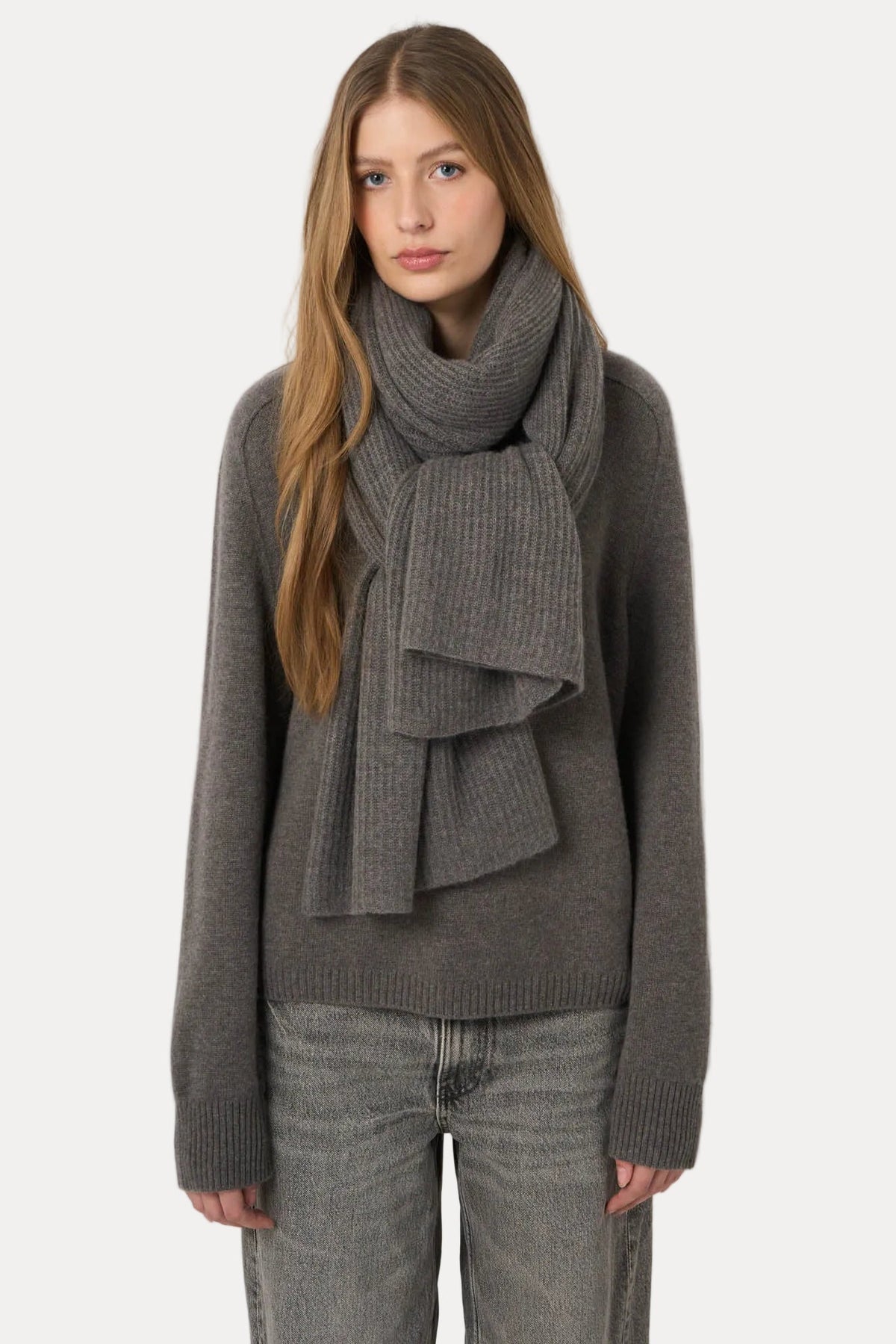 A woman modeling a dark gray ribbed knit sweater and matching scarf against a plain light gray studio background.