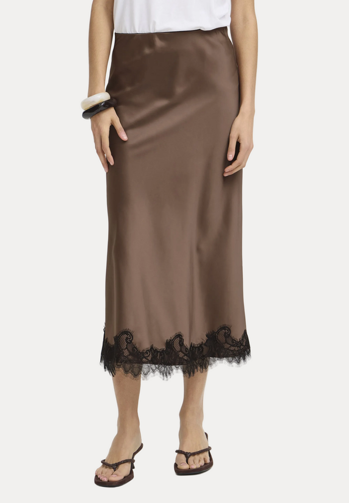A model wearing a brown satin midi skirt with a black lace hem, paired with brown strappy sandals and a white top against a plain white studio background.