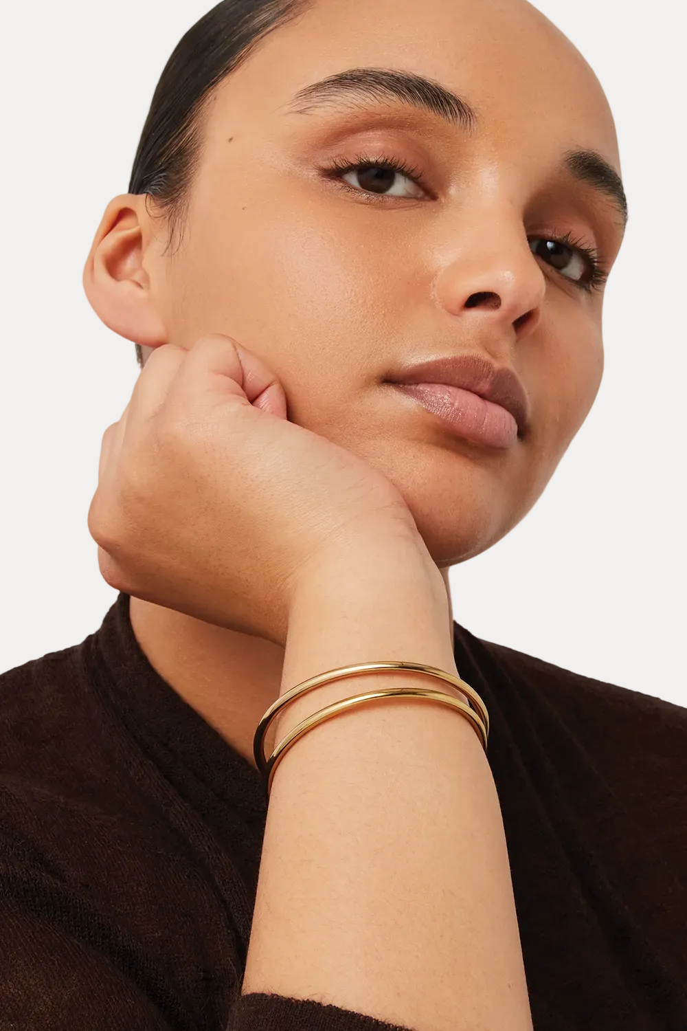 A close-up of a model wearing two slim gold bangle bracelets on her wrist, resting her chin on her hand against a light gray background.
