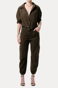 A model wearing an olive brown utility jumpsuit with a button-down shirt collar, rolled sleeves, cargo pockets, and a matching belt, paired with beige high-heeled sandals against a light gray studio background.