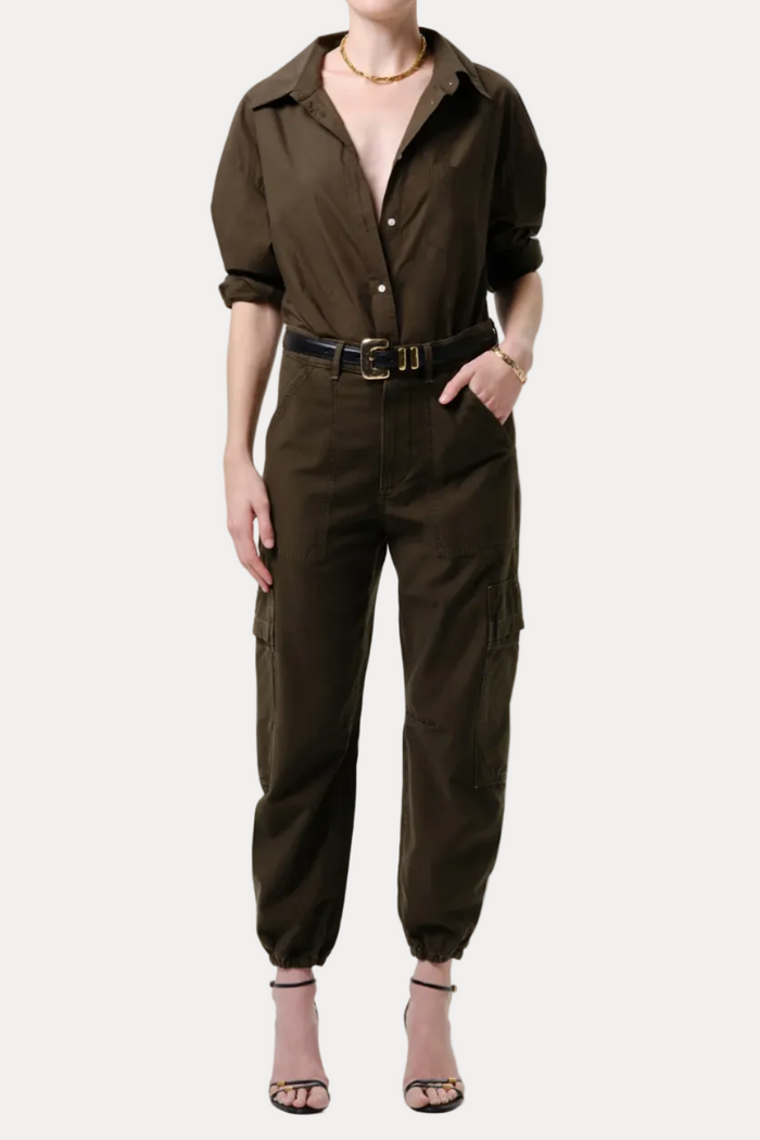 A model wearing an olive brown utility jumpsuit with a button-down shirt collar, rolled sleeves, cargo pockets, and a matching belt, paired with beige high-heeled sandals against a light gray studio background.