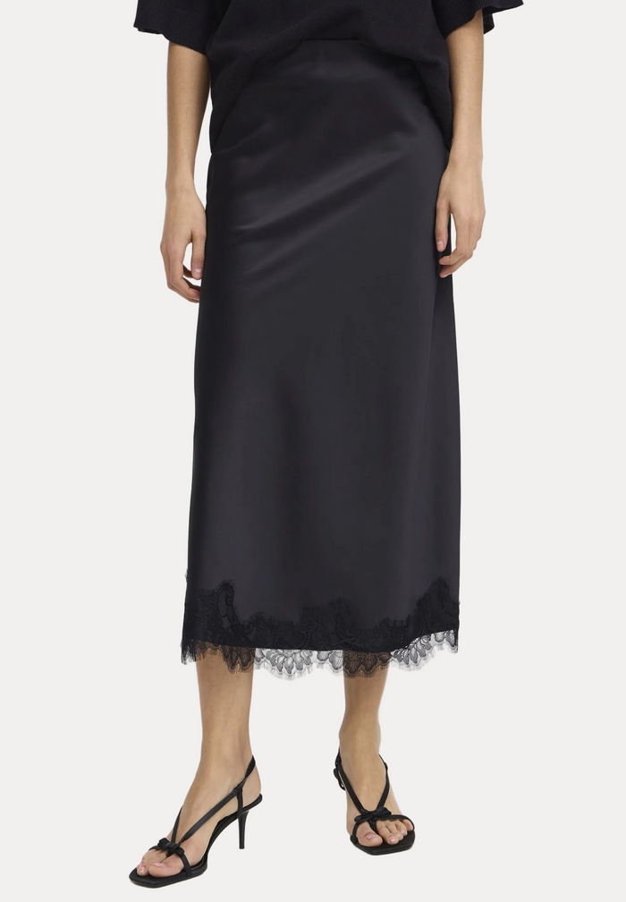 A model wearing a black satin midi skirt with a lace hem, paired with black strappy high-heeled sandals, shown from the waist down on a light gray studio background.
