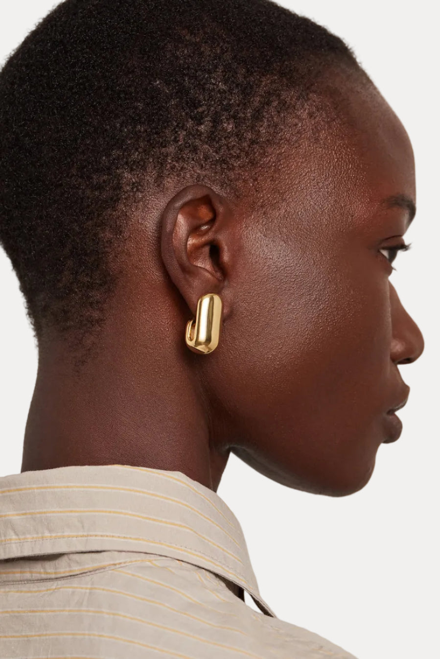 A close-up side profile of a model wearing a chunky, polished gold geometric hoop earring and a beige striped collared shirt against a light gray background.