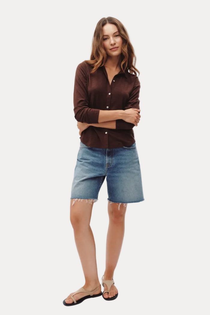 A woman modeling a dark brown long-sleeve button-up shirt and light blue denim shorts with frayed hems, paired with black flat sandals, against a plain white studio background.