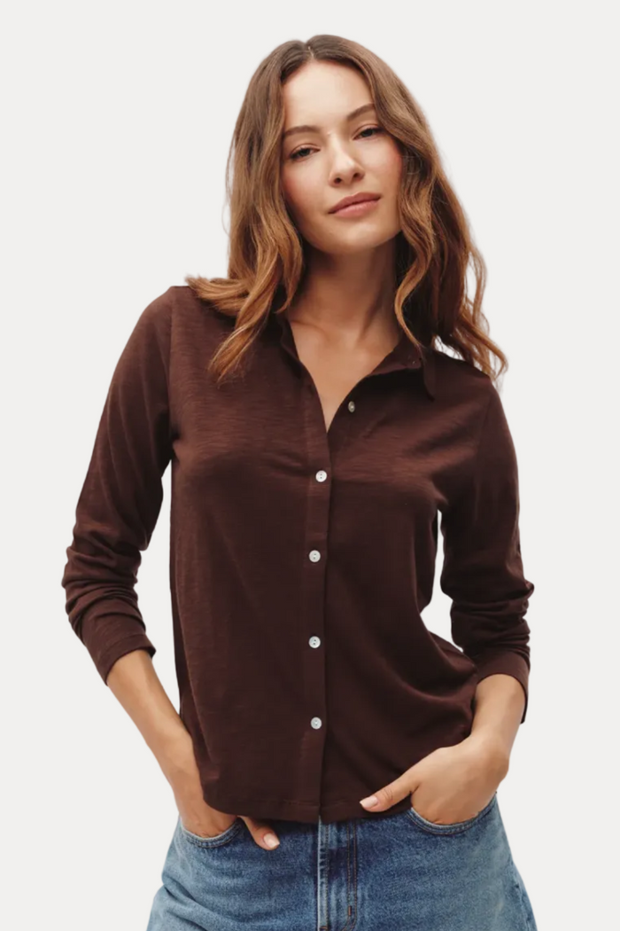 A woman wearing a dark brown long-sleeve button-up shirt and blue jeans, standing against a light gray studio background.