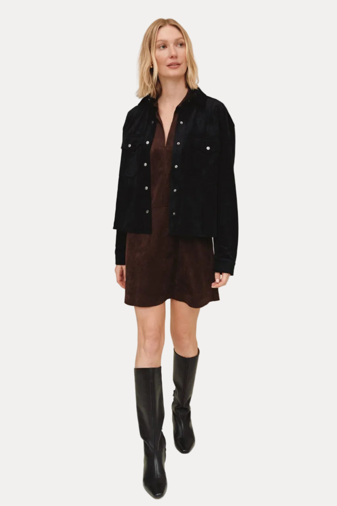 Woman wearing a black jacket over a brown dress with black knee-high boots on a beige background