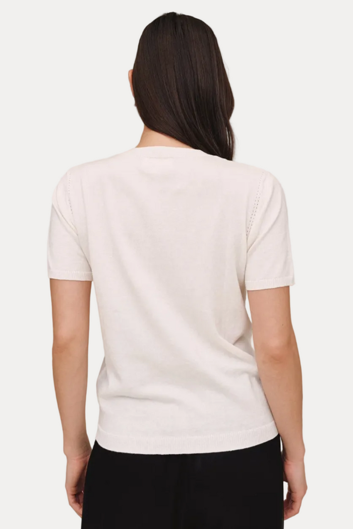 A woman viewed from the back wearing a short-sleeve white knit top and black pants, standing against a plain beige studio background.
