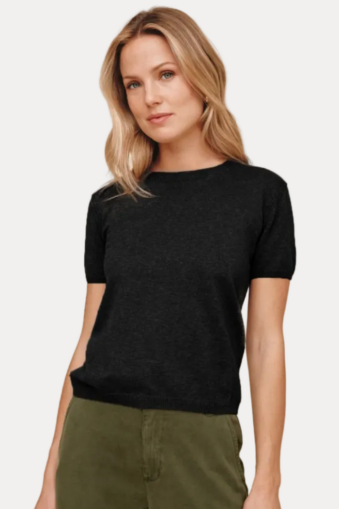 A woman modeling a short-sleeve black knit top with olive green pants, standing against a solid beige studio background.