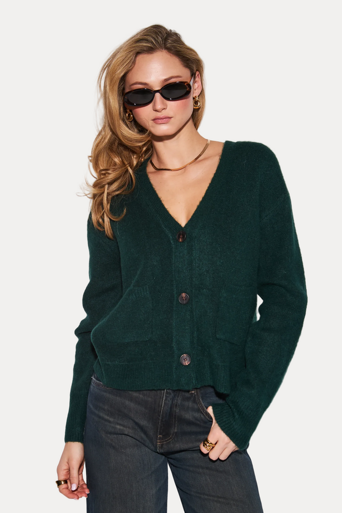 A woman modeling a dark forest green knit cardigan sweater with front buttons, paired with dark wide-leg jeans, sunglasses, and gold jewelry against a plain light gray studio background.