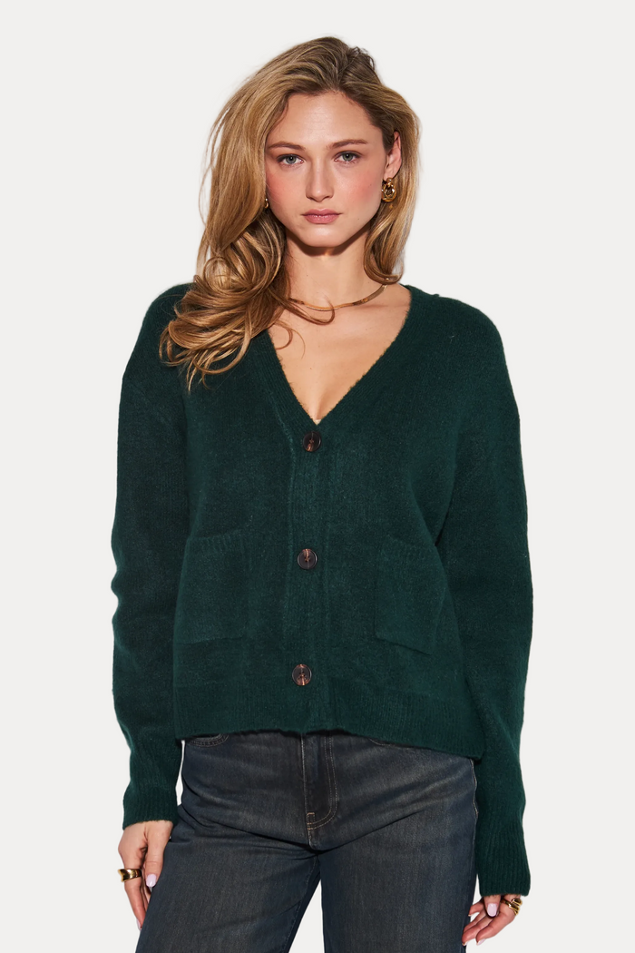 A woman modeling a dark forest green knit cardigan with a V-neck and three front buttons, paired with dark gray jeans, against a plain light gray studio background.