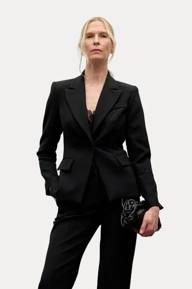 A model stands in a minimalist studio setting wearing a black tailored blazer and matching wide-leg trousers, paired with black high heels and holding a small black clutch.