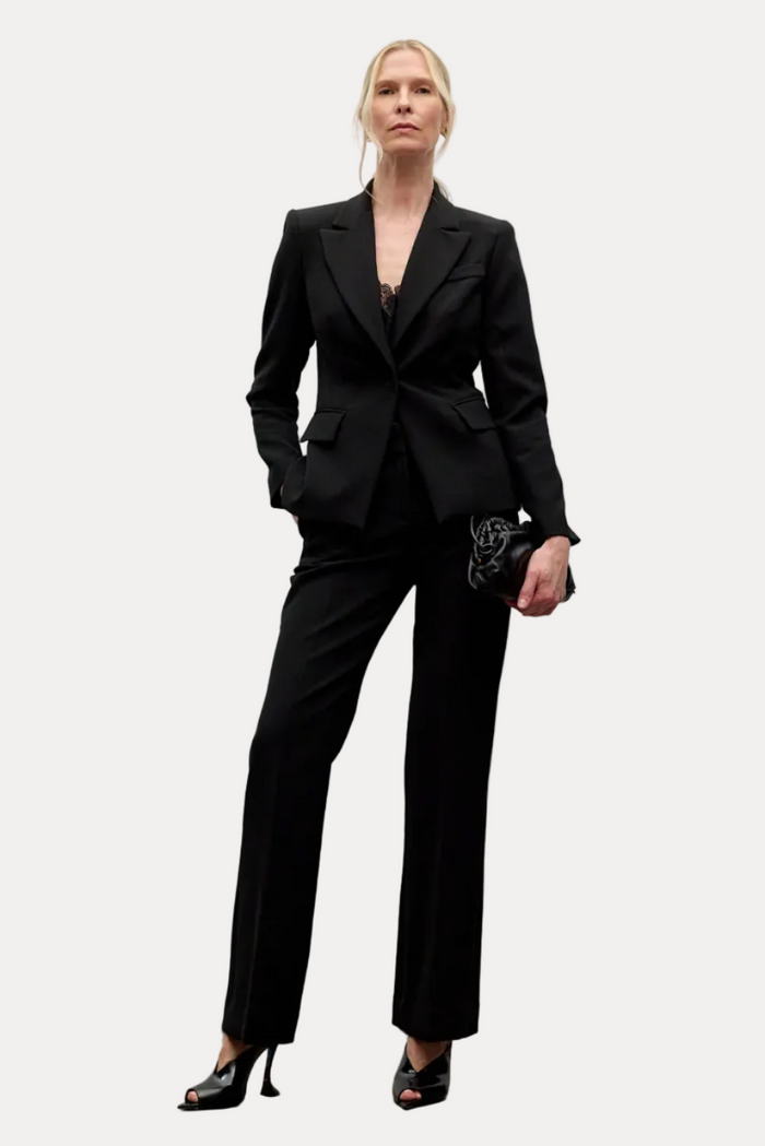 A model stands in a minimalist studio setting wearing a black tailored blazer and matching wide-leg trousers, paired with black high heels and holding a small black clutch.
