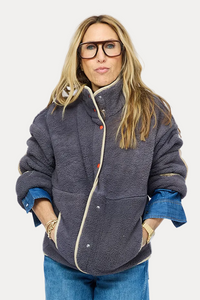 A woman stands against a light gray studio background wearing a dark gray zip-front jacket with blue denim sleeves, blue wide-leg jeans, and brown lace-up shoes.