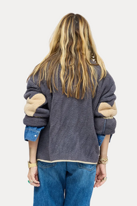 A woman viewed from behind wearing a dark gray patchwork knit sweater with beige elbow patches, wide-leg blue jeans, and brown boots, standing against a plain white studio background.