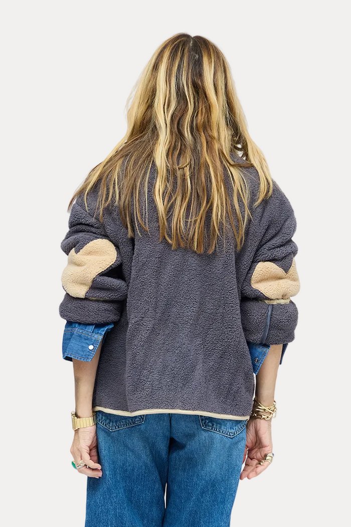 A woman viewed from behind wearing a dark gray patchwork knit sweater with beige elbow patches, wide-leg blue jeans, and brown boots, standing against a plain white studio background.
