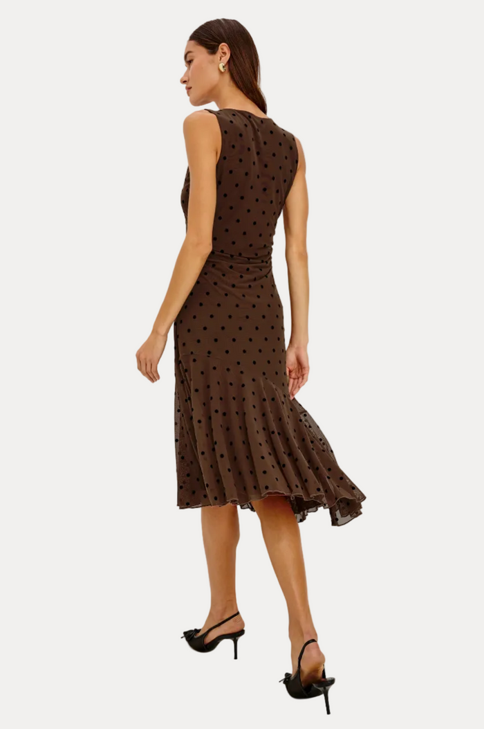 A woman modeling a sleeveless brown midi dress with black polka dots, shown from a rear three-quarter angle against a plain white studio background.