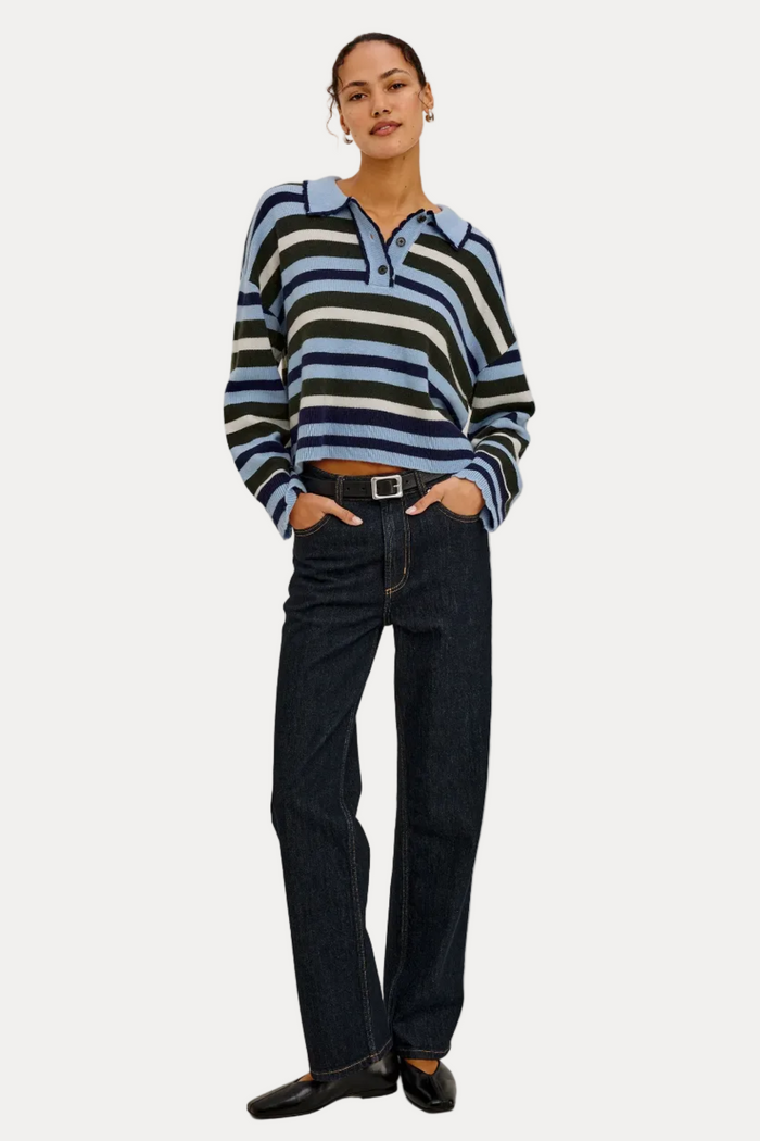 A model stands against a plain light gray studio background wearing a long-sleeve collared knit top with horizontal navy, light blue, black, and white stripes, paired with dark blue jeans and black shoes.