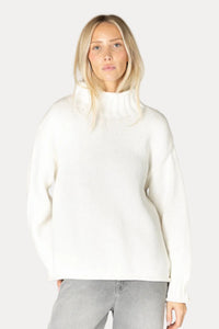 A blonde woman wearing a cream-colored ribbed knit mock neck sweater and light gray jeans, standing against a plain light beige studio background.