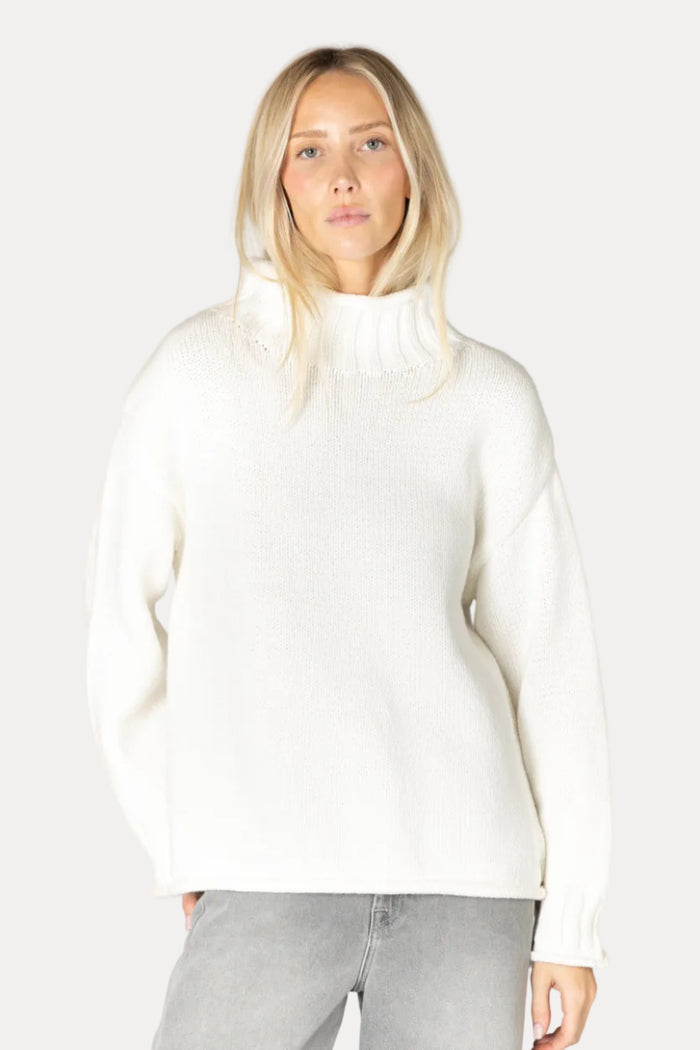 A blonde woman wearing a cream-colored ribbed knit mock neck sweater and light gray jeans, standing against a plain light beige studio background.