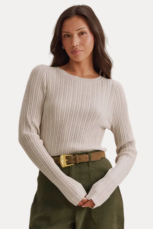 A woman wearing a ribbed cream long-sleeve top and olive green pants with a brown belt, standing against a textured beige background.