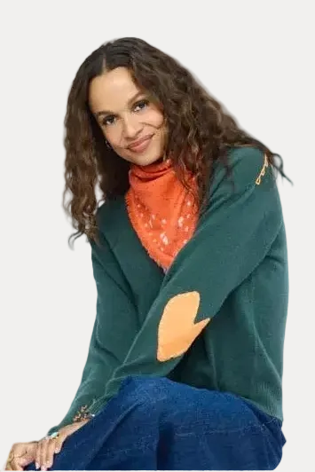 A woman with long curly brown hair is seated against a plain white background, wearing a dark green knit sweater with a tan elbow patch, an orange patterned turtleneck, and blue jeans.