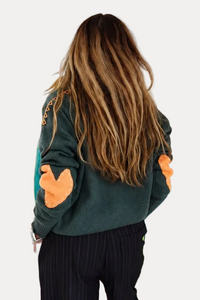 A woman with long light brown hair is seen from behind, wearing a dark green sweatshirt with orange patches on the sleeves and black pinstripe trousers, standing in a room with colorful abstract paintings on the walls.