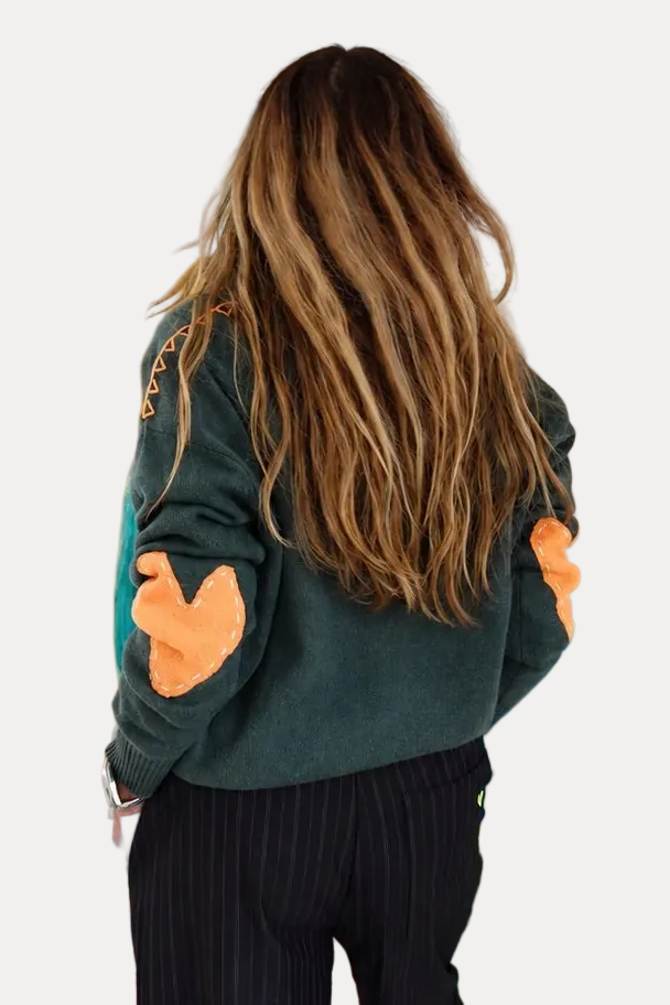 A woman with long light brown hair is seen from behind, wearing a dark green sweatshirt with orange patches on the sleeves and black pinstripe trousers, standing in a room with colorful abstract paintings on the walls.