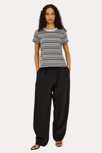 A model stands against a plain light gray studio background wearing a short-sleeve crew-neck t-shirt with thin horizontal navy, gray, and white stripes, paired with loose black drawstring pants and black sandals.