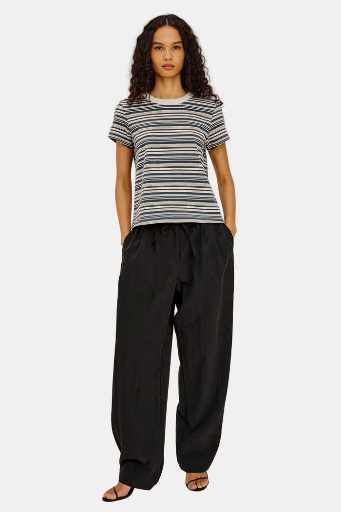 A model stands against a plain light gray studio background wearing a short-sleeve crew-neck t-shirt with thin horizontal navy, gray, and white stripes, paired with loose black drawstring pants and black sandals.