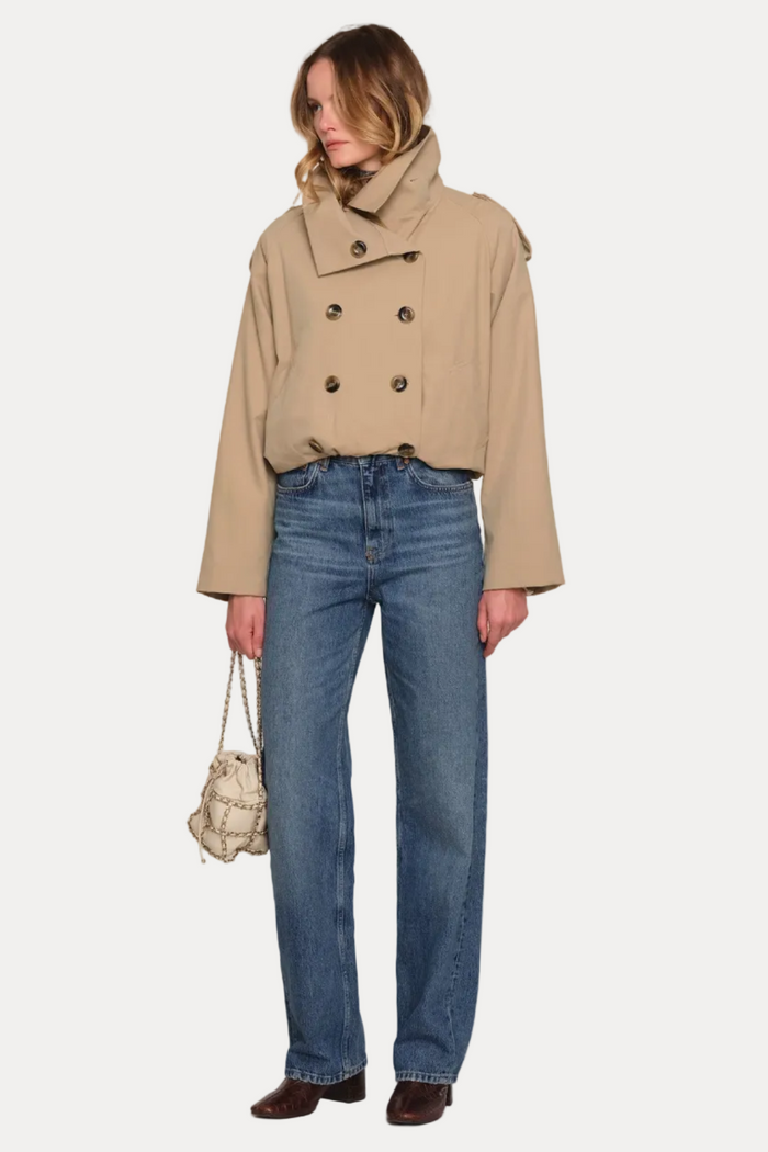 A model stands against a plain light beige studio background wearing a cropped beige double-breasted trench jacket, blue straight-leg jeans, dark brown shoes, and holding a small woven cream handbag.