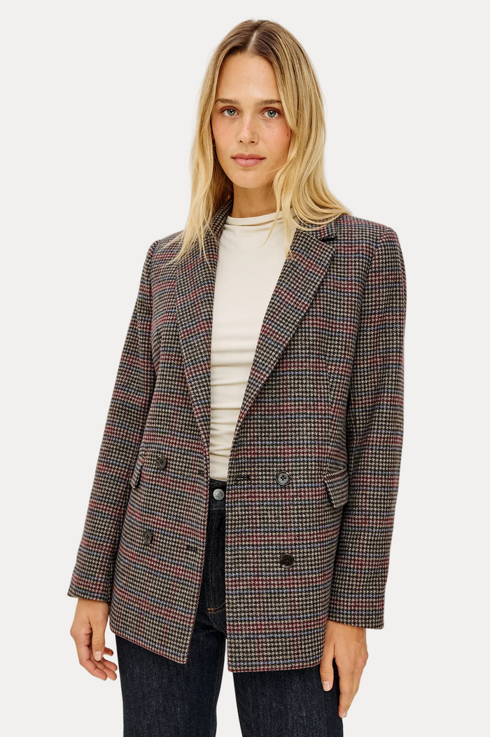 A woman modeling a double-breasted blazer with a fine black, red, and beige houndstooth pattern, worn over a cream ribbed top and dark jeans, against a plain light gray background.