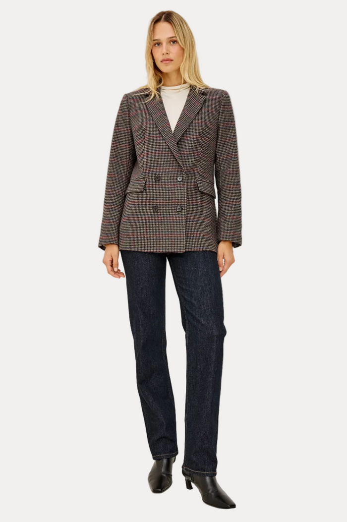 A woman modeling a double-breasted blazer in a brown and gray micro-check pattern, paired with dark blue jeans and black ankle boots, against a plain white studio background.