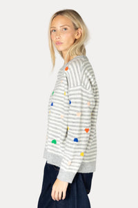 A woman wearing a gray and white striped knit sweater with colorful heart-shaped pom-poms, paired with dark blue pants, photographed against a plain light background.