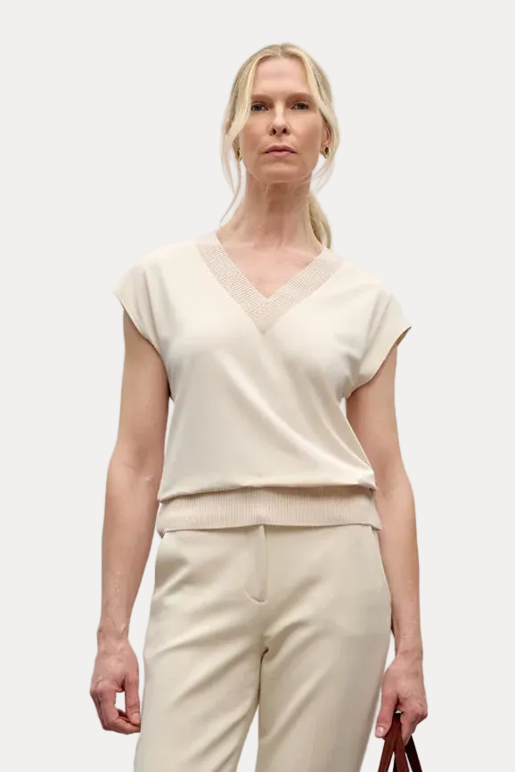 A woman stands against a plain light gray wall wearing a cream short-sleeve V-neck top and matching cream wide-leg trousers, holding a small brown handbag.