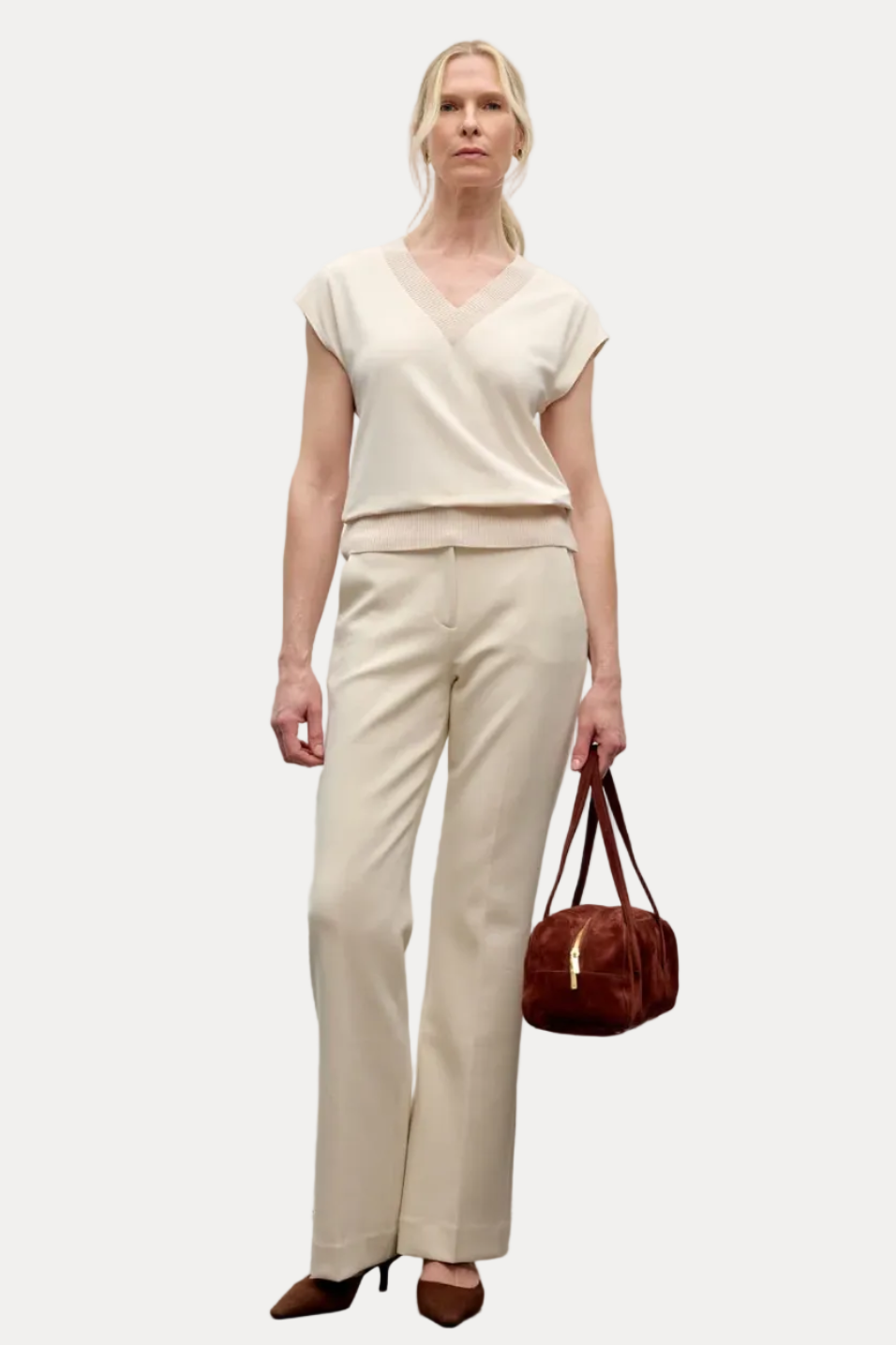 A woman stands against a plain light gray wall wearing a cream short-sleeve V-neck top and matching cream wide-leg trousers, holding a small brown handbag.